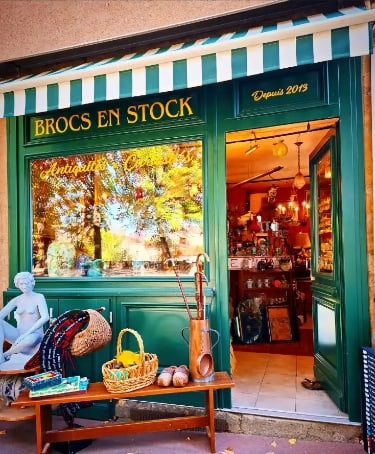 La devanture verte de Brocs en Stock sous son store rayé, une statue et des paniers sur le trottoir