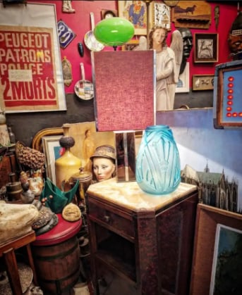 Un coin de la boutique : tableaux, affiche ancienne, vase en verre et lampe sur une table de chevet