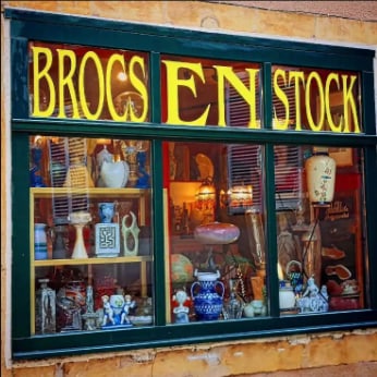 Une vitrine aux grandes lettres jaunes « Brocs en Stock », remplie de céramiques et de lampes